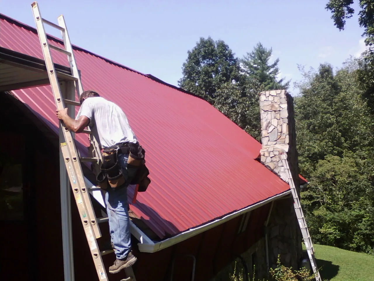 Roofer on a ladder beside a red metal roof and stone chimney — Soffit & Fascia Repair by Roofing Brothers in Lengby